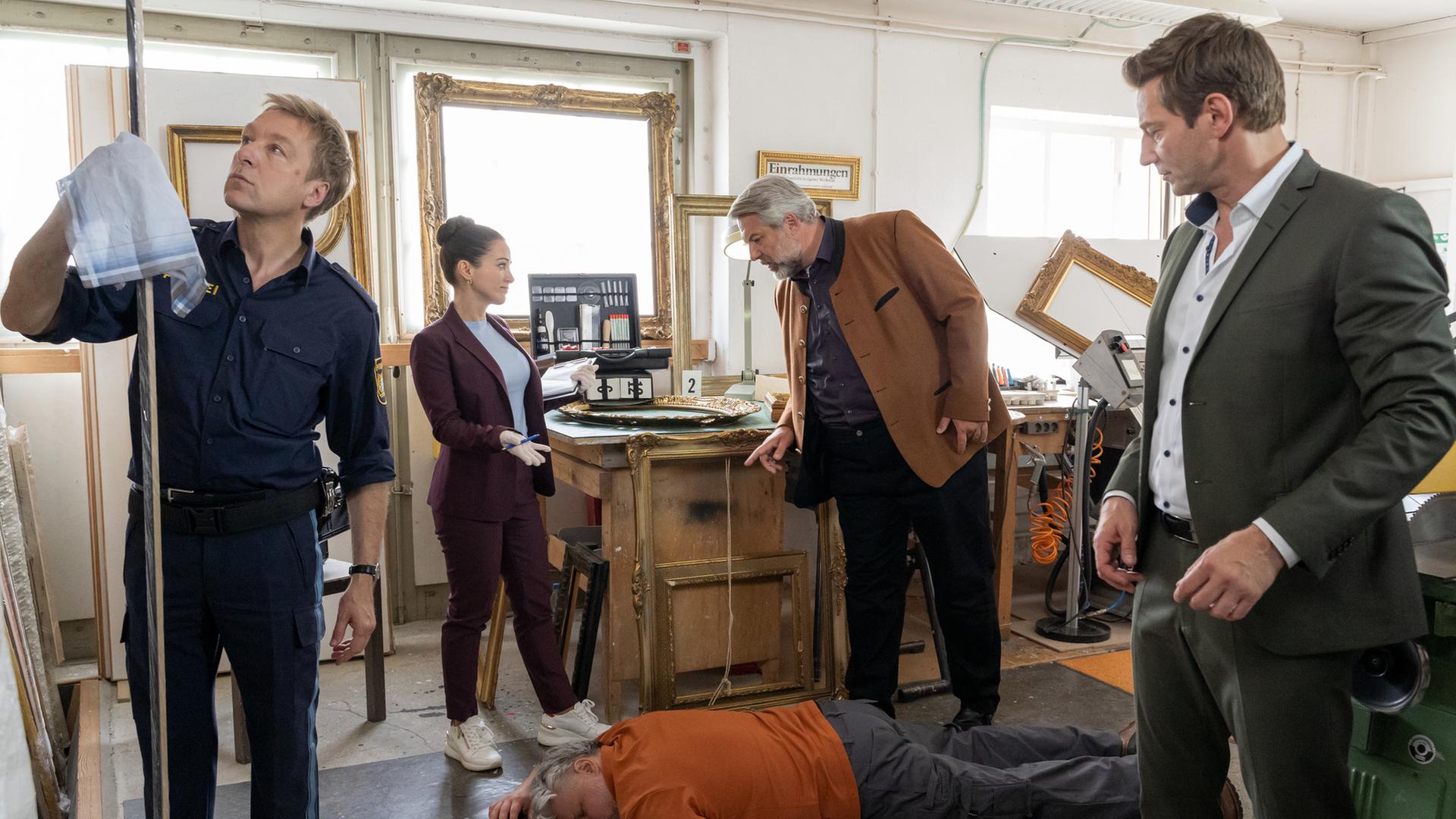 "Die Rosenheim-Cops - Ein grober Schnitzer": Polizeihauptmeister Mohr (Max Müller), Rechtsmedizinerin Ela Atay (Sevda Polat), Kommissar Stadler (Dieter Fischer) und Kommissar Hansen (Igor Jeftić) untersuchen den Tatort um das Opfer Stefan Ertl (Komparse), das am Boden liegt. Mohr hält mit einem Tuch eine Rahmenleiste fest, im Hintergrund sind diverse goldene Bilderrahmen zu sehen.
