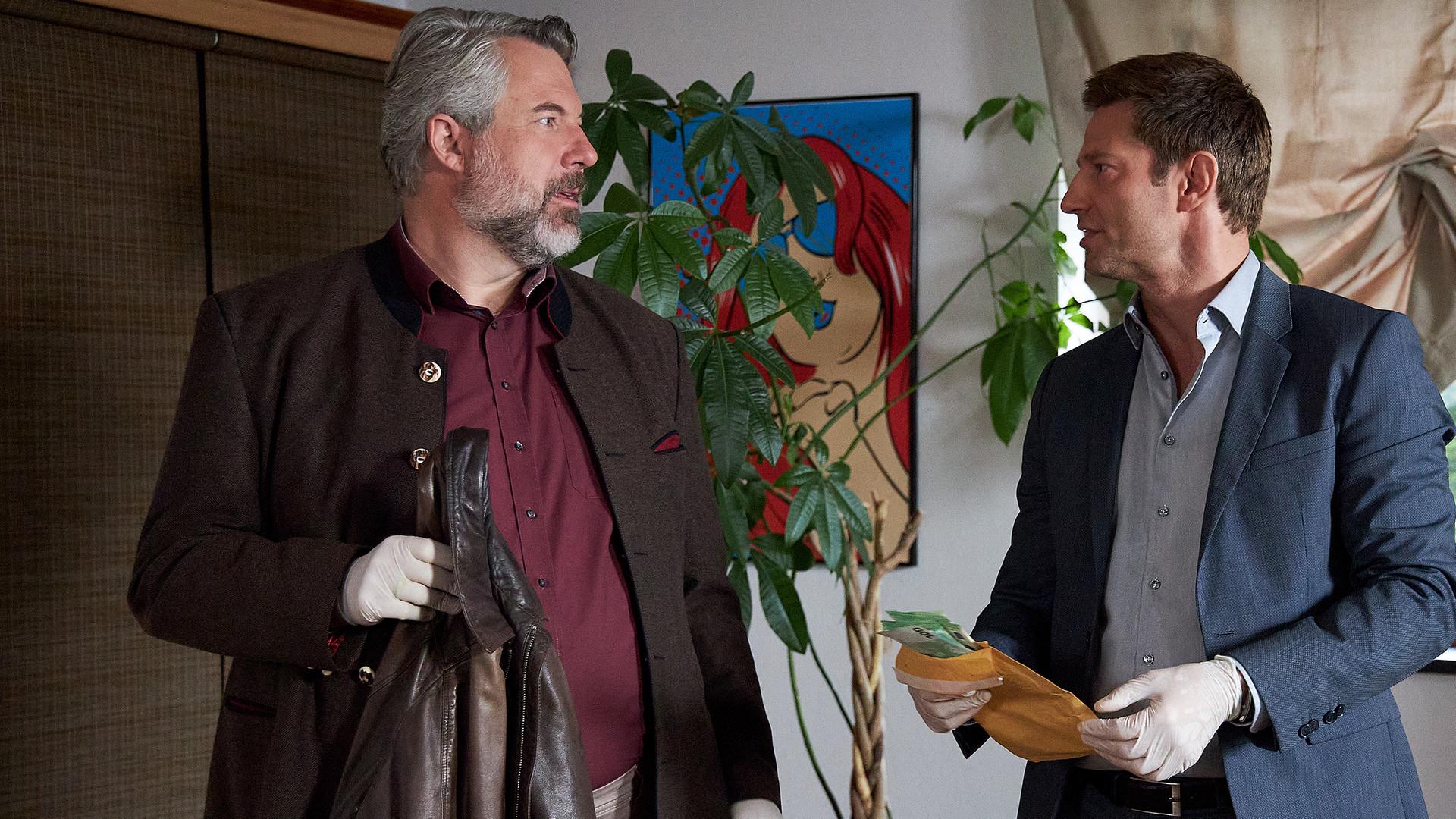 Die Kommissare Stadler (Dieter Fischer) und Hansen (Igor Jeftić) stehen in einer Wohnung und sehen sich an. Sie tragen weiße Schutzhandschuhe. Stadler hält eine dunkelbrauen Lederjacke in der Hand, Hansen einen Umschlag mit Geldscheinen. Im Hintergrund sind eine große Topfpflanze zu sehen sowie ein modernes Bild neben einem Fenster, das von oben herab halb mit einem Vorhang abgedeckt ist.