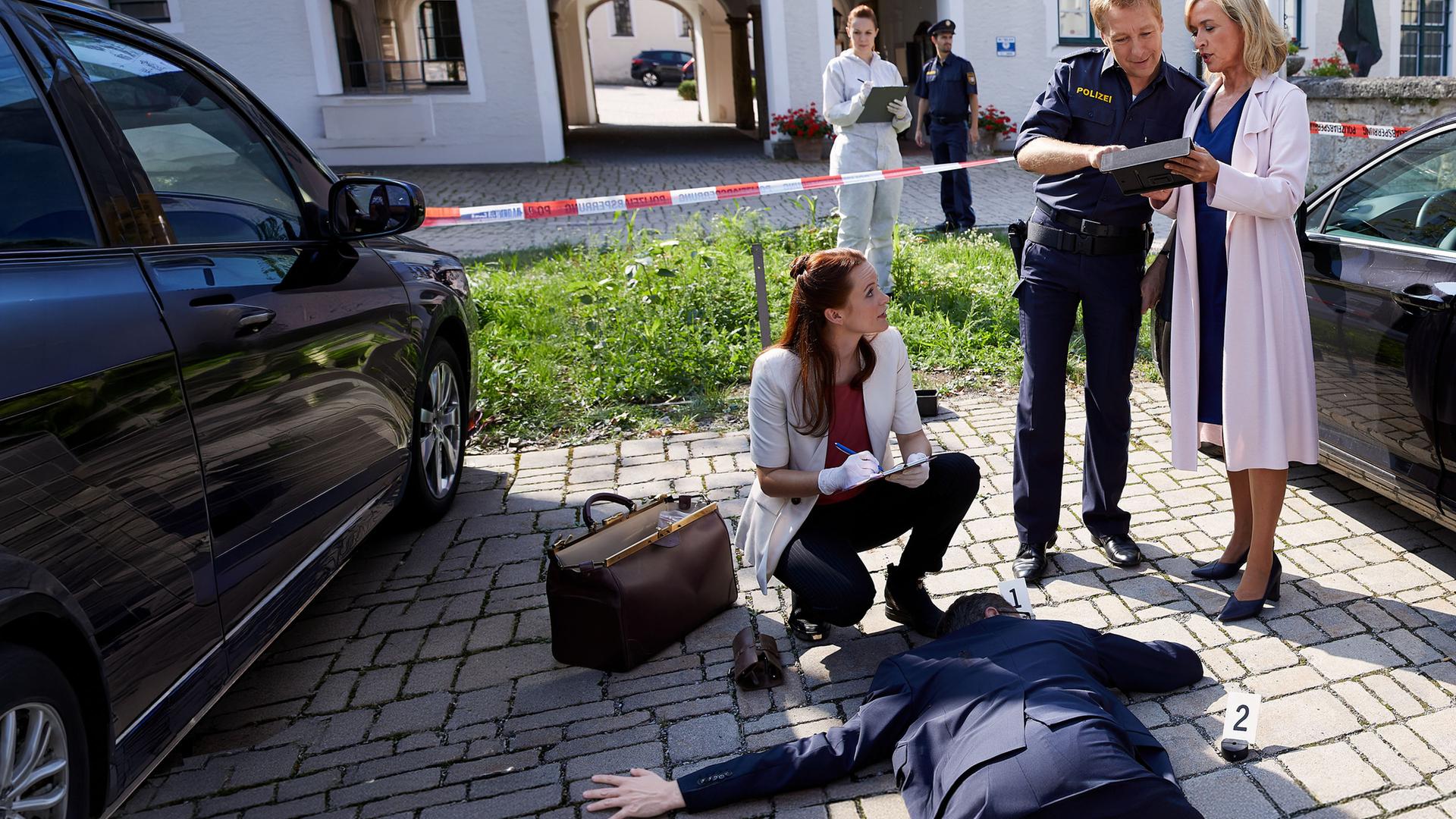 "Die Rosenheim-Cops - Ein Haus für alle Fälle": Pathologin Mai (Sina Wilke) kniet vor dem Opfer, das neben einem schwarzen Auto bäuchlings auf dem Parkplatz liegt. Daneben stehen Polizeihauptmeister Mohr (Max Müller) und Kommissarin Danner (Katharina Abt), die gemeinsam auf ein Tablet schauen. Im Hintergrund Kollegen der Polizei.