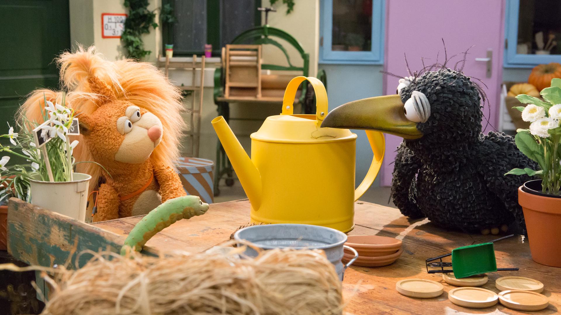 Eichhörnchen Ella und Rabe Rudi schauen sich gespannt an. Zwischen ihnen steht eine große, gelbe Gießkanne, vor ihnen hockt Wurm Lotti.