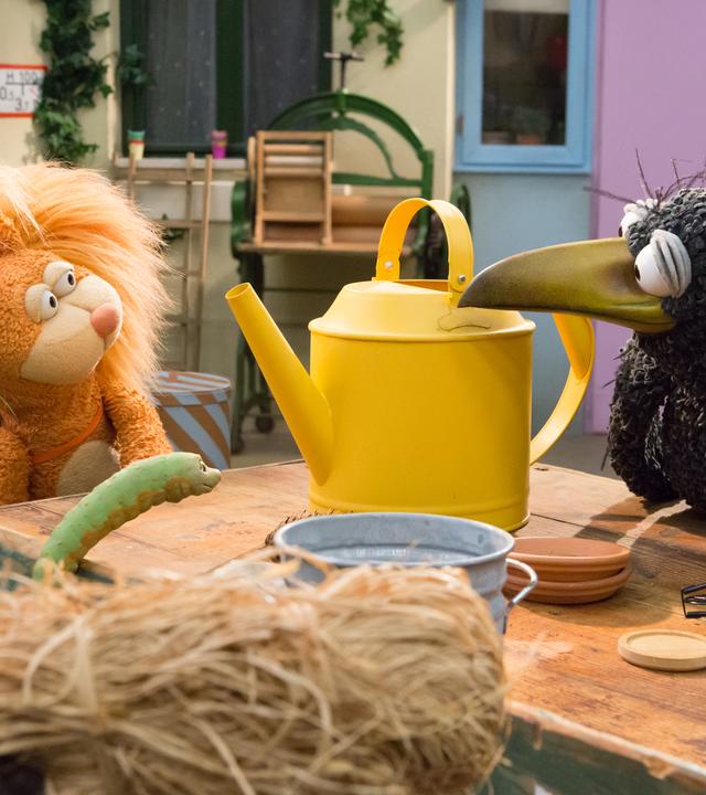 Eichhörnchen Ella und Rabe Rudi schauen sich gespannt an. Zwischen ihnen steht eine große, gelbe Gießkanne, vor ihnen hockt Wurm Lotti.