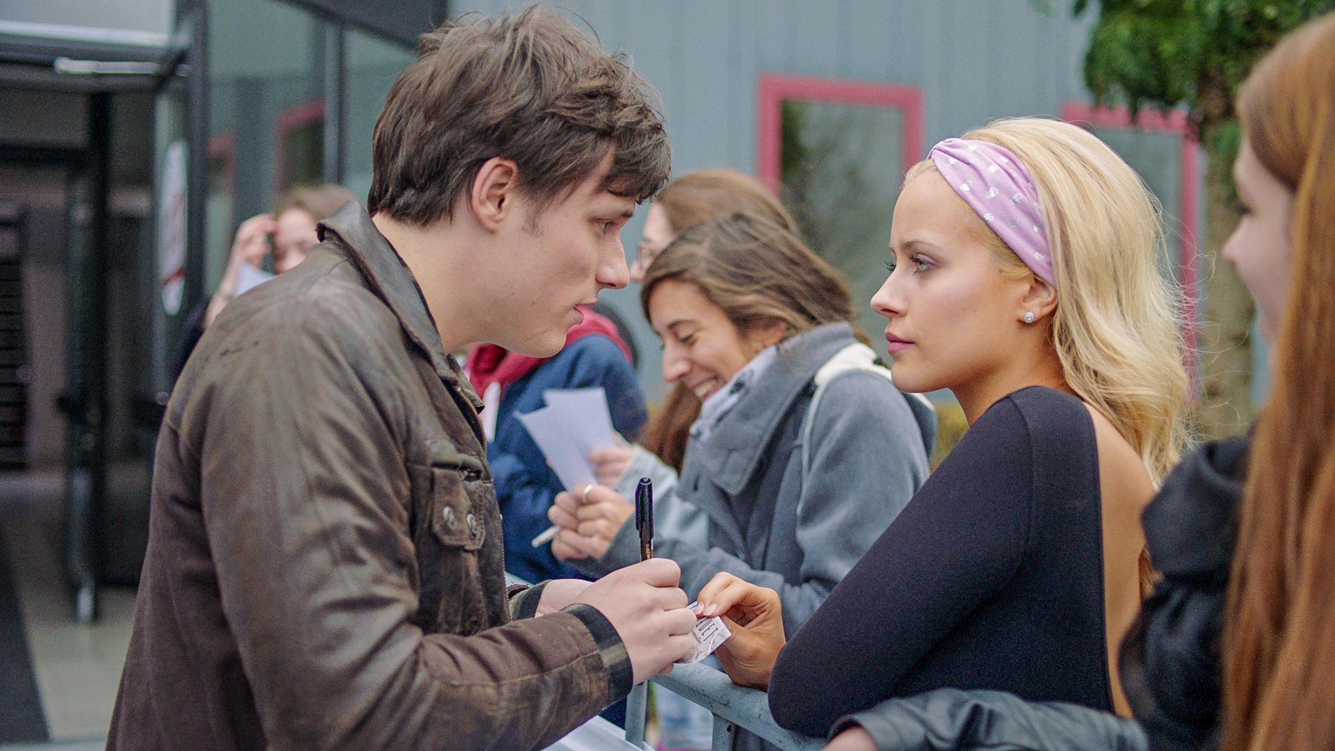 Lena steht mit Henris Fans am Radiosender. Henri unterschreibt eine Autogrammkarte für sie. Lena wirkt wütend.