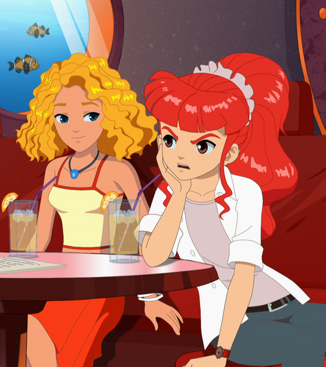 Rikki, Emma und Cleo sitzen zusammen im Café. Rikki schaut sehr verärgert.