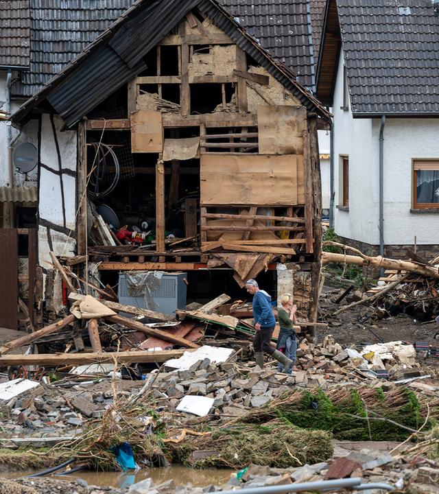 Im Hintergrund zerstörte Häuser, im Vordergrund Trümmer auf dem Boden. Dazwischen einige Menschen, die versuchen aufzuräumen.