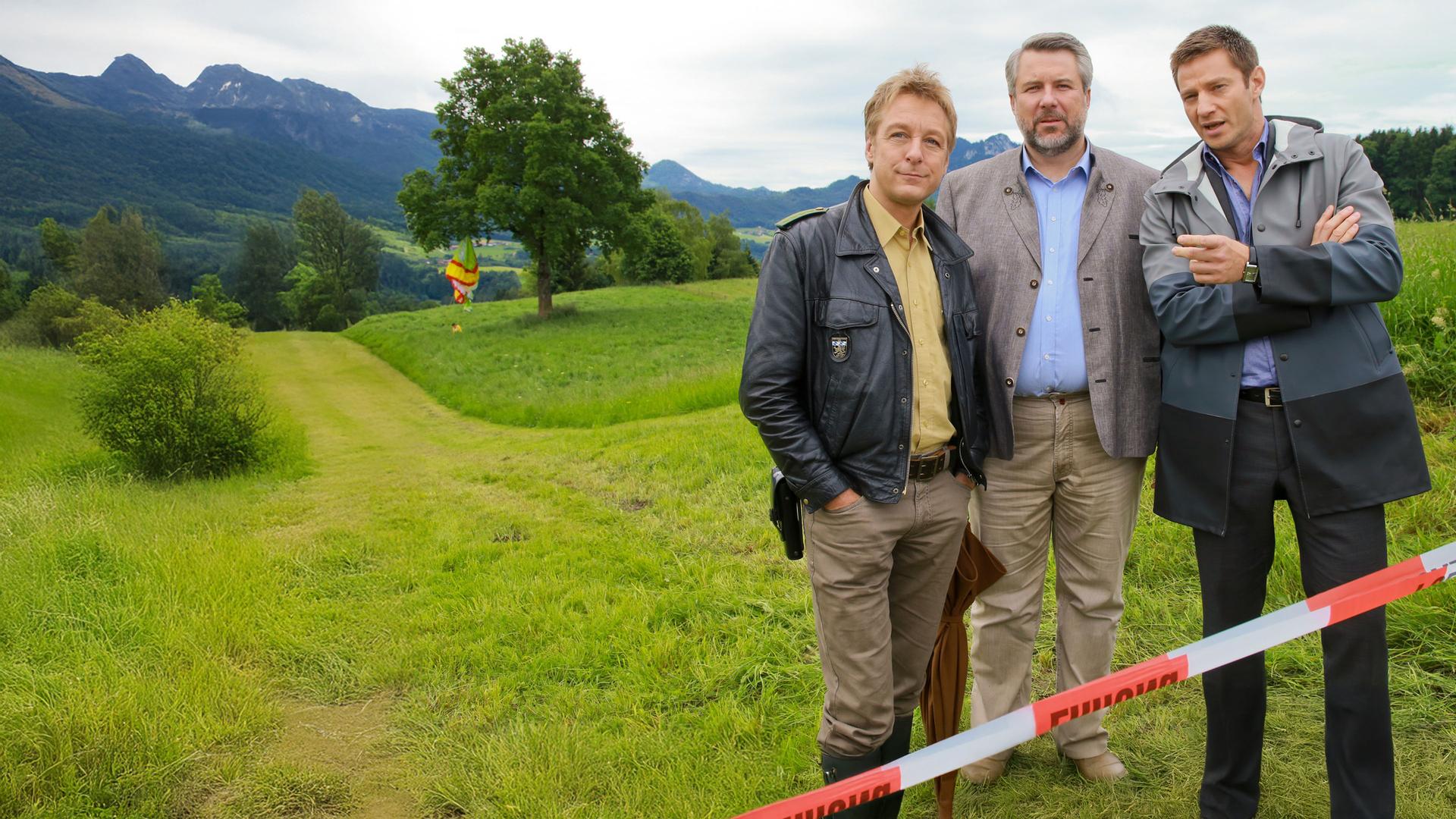 "Die Rosenheim-Cops - Ein Karton kommt selten allein": Auf einer Wiese stehen die Kommissare Stadler (Dieter Fischer), Hansen (Igor Jeftic) und Polizist Mohr (Max Müller). Im Hintergrund befindet sich ein Baum, in dem sich ein Gleitschirm verfangen hat.