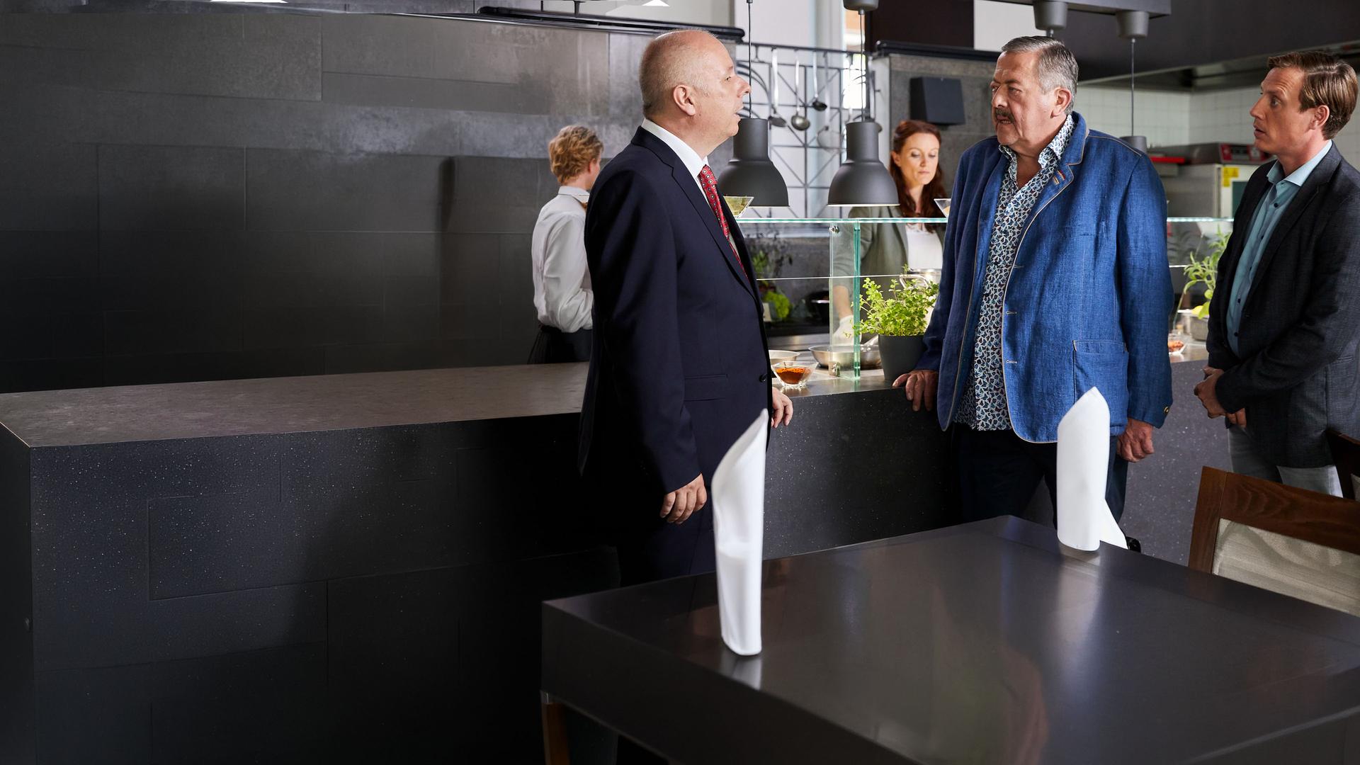 "Die Rosenheim-Cops - Ein Koch zu viel": Die Kommissare Hofer (Joseph Hannesschläger) und Bach (Patrick Kalupa) stehen in einem Restaurant Richard Mayerhofer (Markus H. Eberhard) gegenüber und reden. Im Hintergrund untersucht Pathologin Wilke den Tatort.