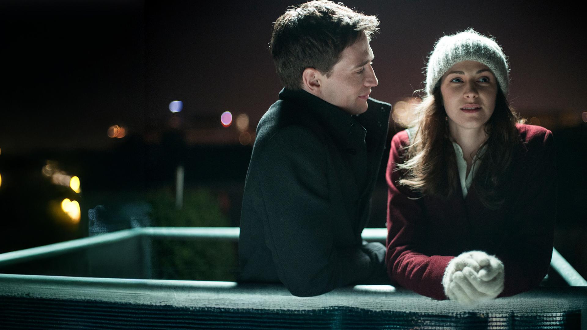 "Ein Lächeln nachts um vier": Max (August Wittgenstein) und Jule (Natalia Belitski) stehen bei Nacht draußen und lehnen sich auf ein Geländer. Sie sind winterlich gekleidet.