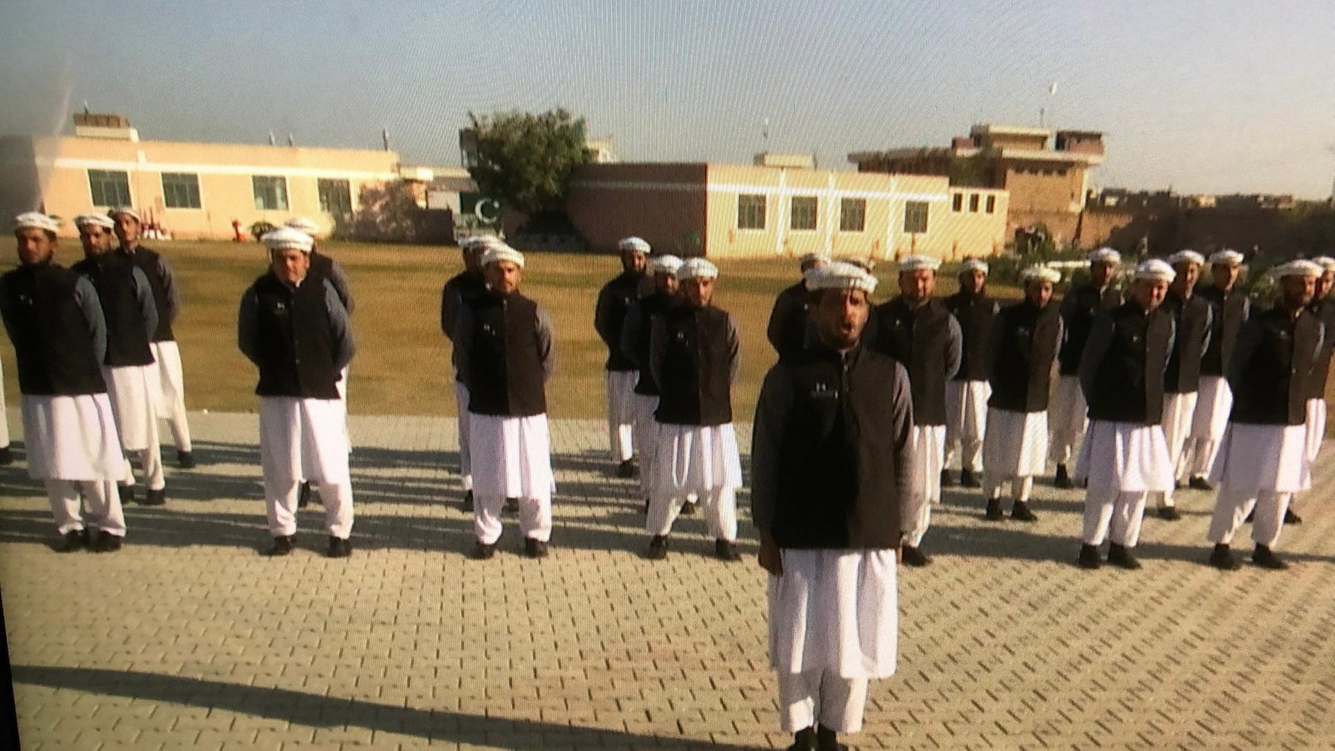 "Ein Leben nach dem Terror – Pakistan und die Ex-Taliban": Mehrere Männer in weißen Hosen, schwarzen Jacken und weißen Hüten stehen in einer Formation und blicken geradeaus.