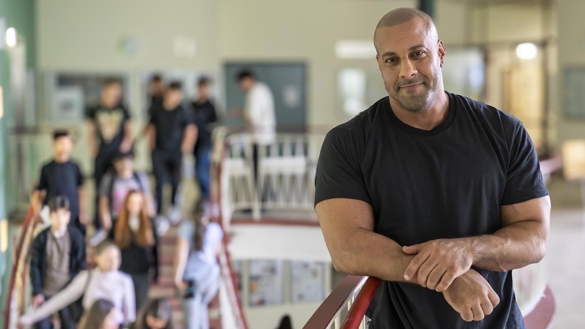 Lehrer Reff lehnt am Treppengeländer in seiner Schule. Auf der Treppe sind mehrere Schüler zu sehen.