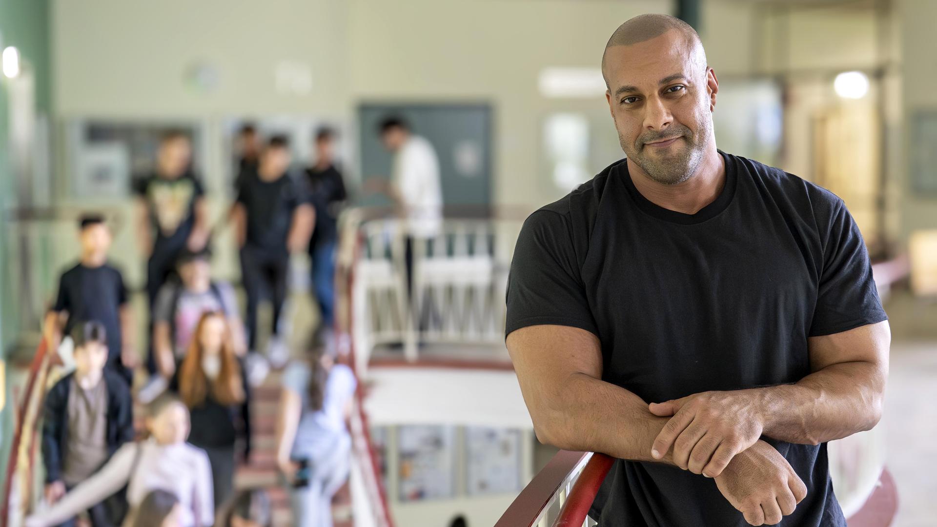 Lehrer Reff lehnt am Treppengeländer in seiner Schule. Auf der Treppe sind mehrere Schüler zu sehen.