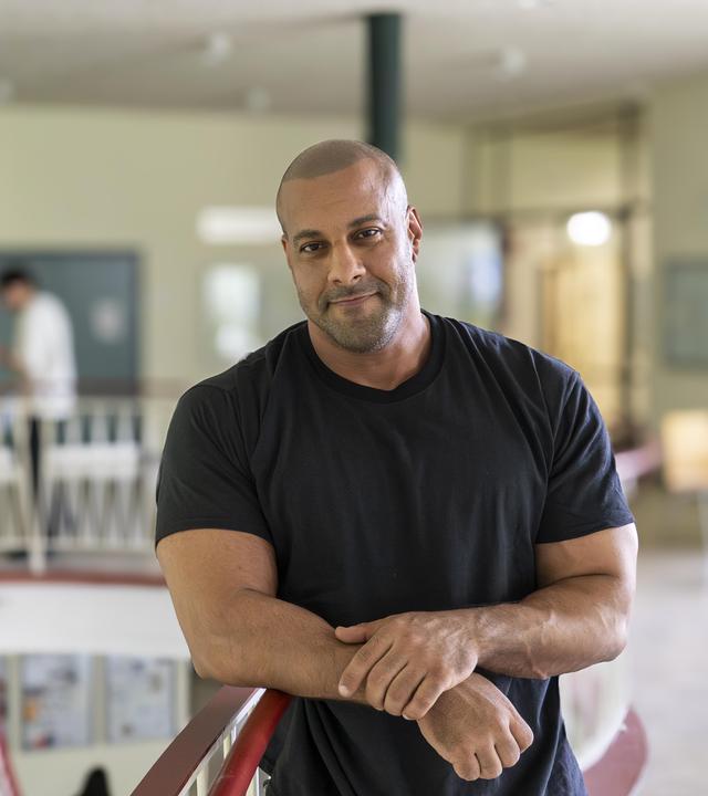 Lehrer Reff lehnt am Treppengeländer in seiner Schule. Auf der Treppe sind mehrere Schüler zu sehen.
