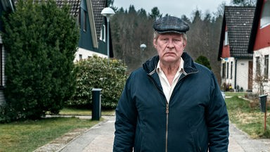 Ove (Rolf Lassgård) steht mit ernstem Gesicht auf einem gepflasterten Weg in seiner Wohnsiedlung.