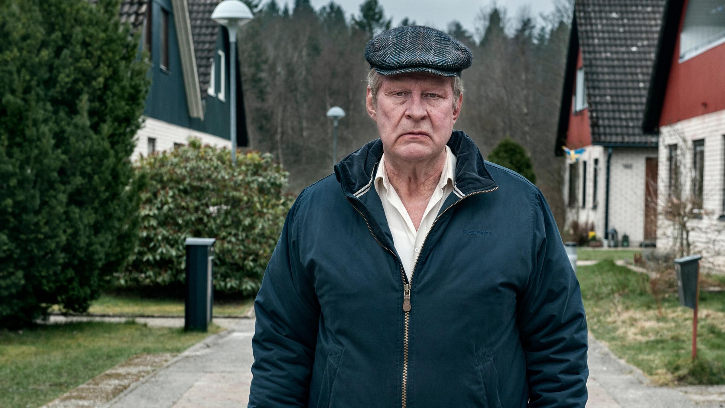 Ove (Rolf Lassgård) steht mit ernstem Gesicht auf einem gepflasterten Weg in seiner Wohnsiedlung.
