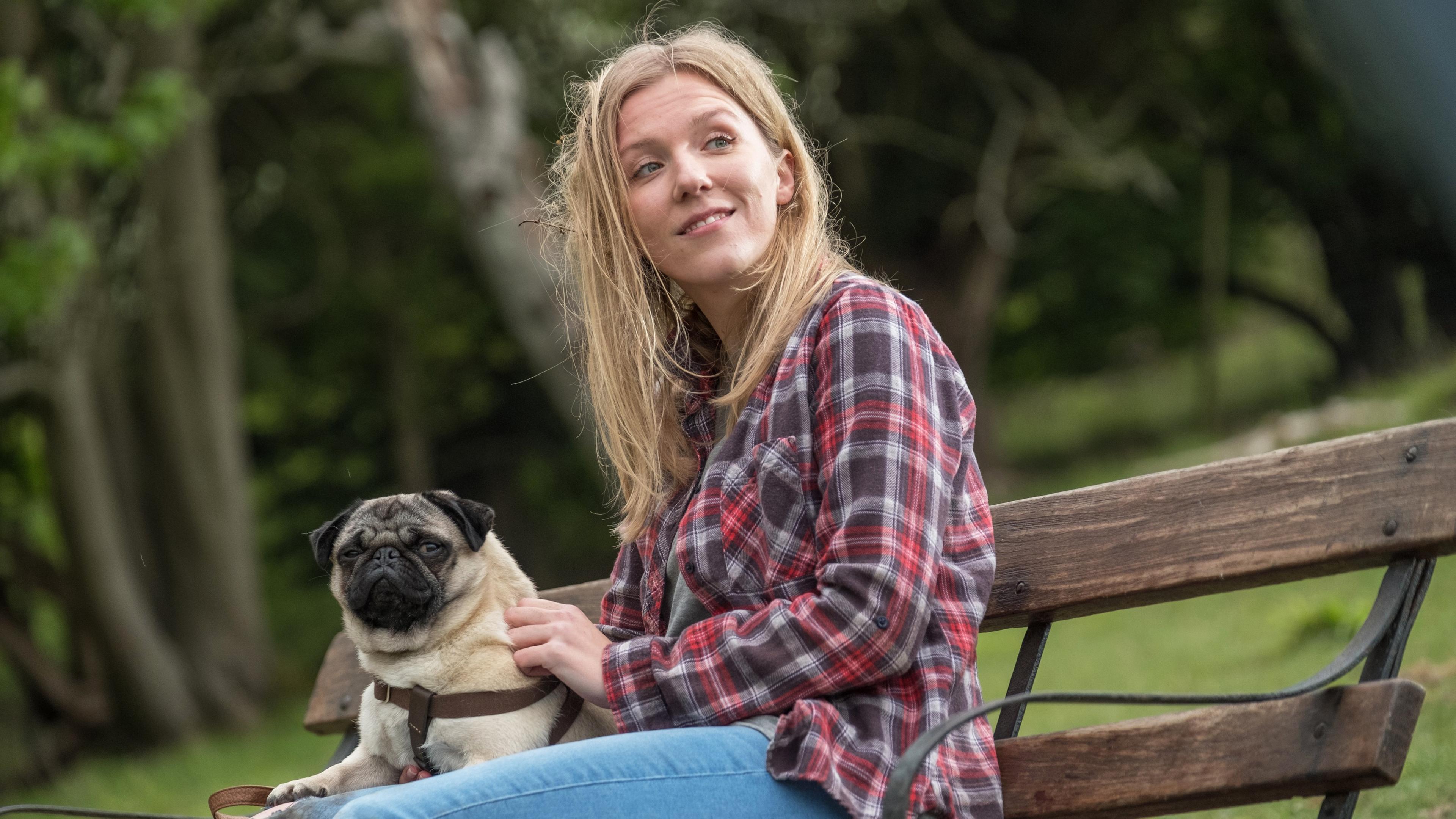 Sarah (Beattie Edmondson) sitzt mit Mops Patrick auf einer Parkbank. Beide schauen interessiert in die gleiche Richtung.