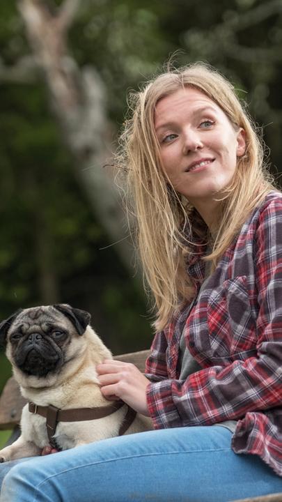 Sarah (Beattie Edmondson) sitzt mit Mops Patrick auf einer Parkbank. Beide schauen interessiert in die gleiche Richtung.