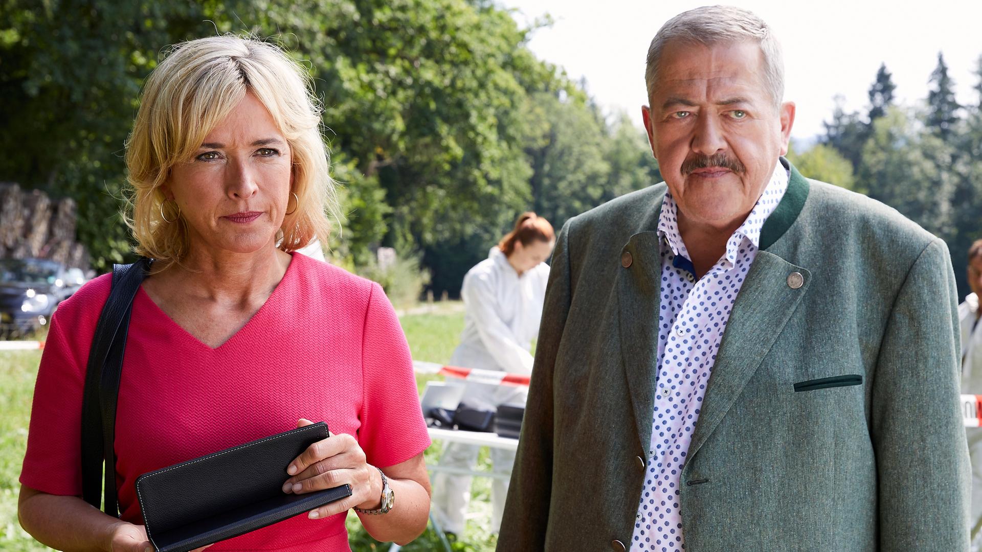 "Die Rosenheim-Cops - Ein mysteriöser Blumenstrauß": Die Kommissare Danner (Katharina Abt) und Hofer (Joseph Hannesschläger) stehen nebeneinander auf einer Wiese und schauen in dieselbe Richtung. Im Hintergrund arbeitet Kollegin (Komparsin) der KTU an einem Tisch hinter rot-weißem Absperrband.