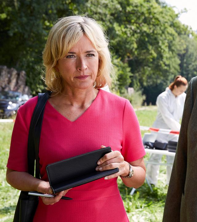 "Die Rosenheim-Cops - Ein mysteriöser Blumenstrauß": Die Kommissare Danner (Katharina Abt) und Hofer (Joseph Hannesschläger) stehen nebeneinander auf einer Wiese und schauen in dieselbe Richtung. Im Hintergrund arbeitet Kollegin (Komparsin) der KTU an einem Tisch hinter rot-weißem Absperrband.