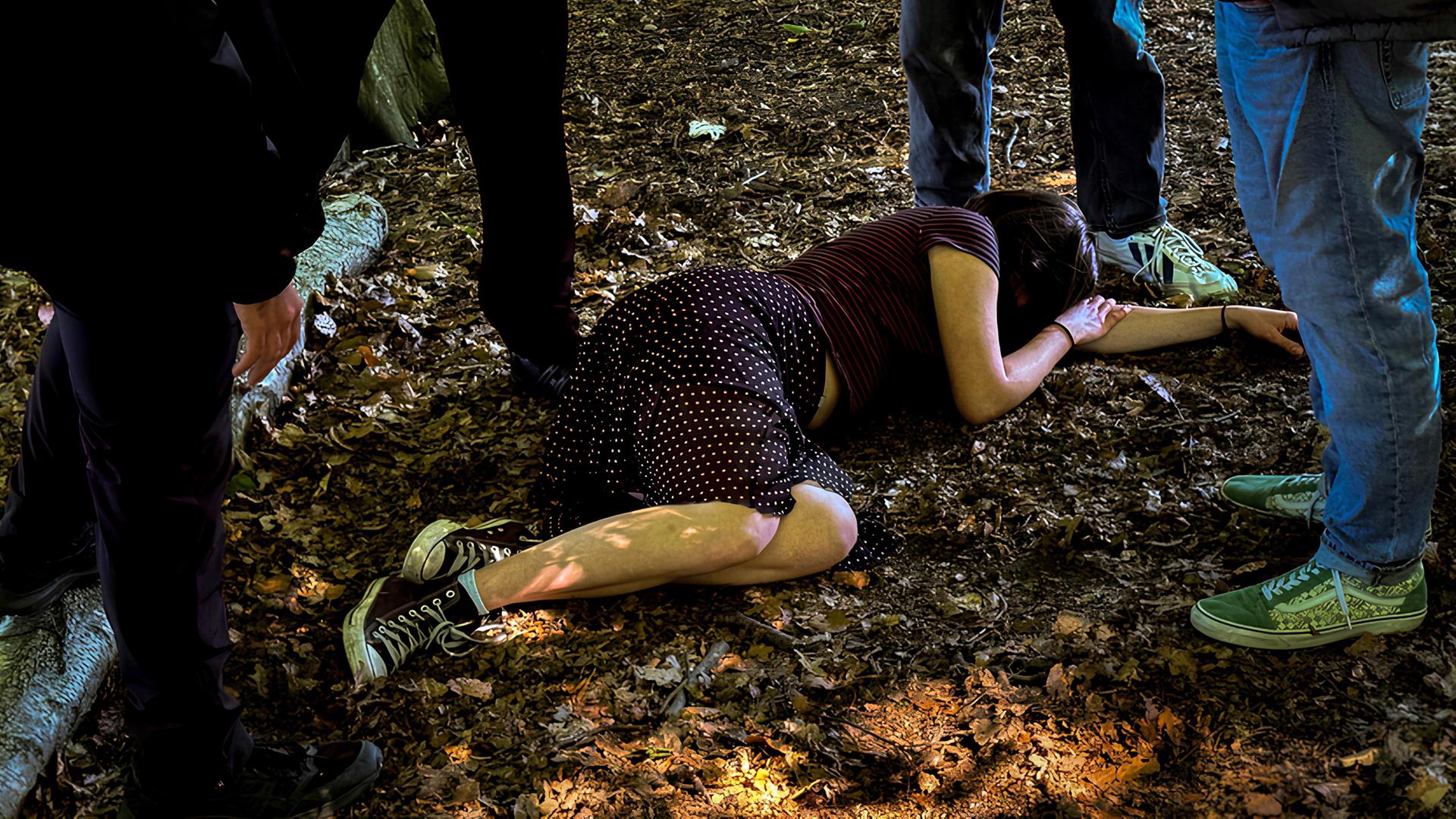 Eine junge Frau liegt auf einem Waldboden, umringt von Männern.