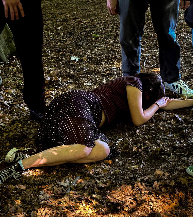 Eine junge Frau liegt auf einem Waldboden, umringt von Männern.