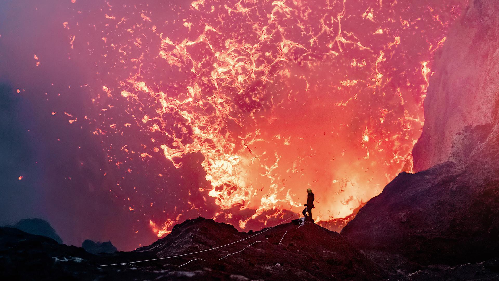 Nahaufnahme eines ausbrechenden Vulkankraters. Am Krater steht eine Person in Schutzkleidung