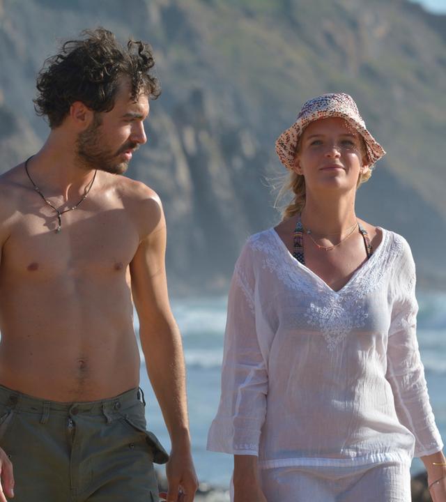"Ein Sommer an der Algarve": An der Meeresküste laufen Miguel Silva (Giovanni Funiati) und Julika Bergmann (Bea Brocks) nebeneinander her. Er schaut zu ihr und sie schaut nach vorne.