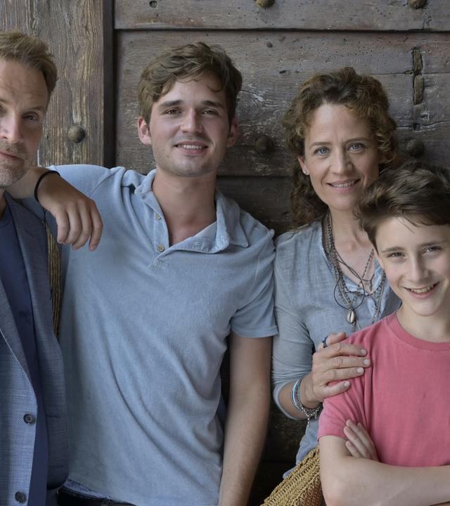 "Ein Sommer auf Elba": Vor einer dunklen Tür auf einer Terrasse steht Maja und ihre Familie. Von links: ihr Ex-Mann Thorsten (Janek Rieke), daneben ihr 17-jähriger Sohn Anton (Henry Horn). Maja (Regula Grauwiller) hat ihrem jüngeren Sohn Erik (Levis Kachel) liebevoll die Hände auf die Schultern gelegt.