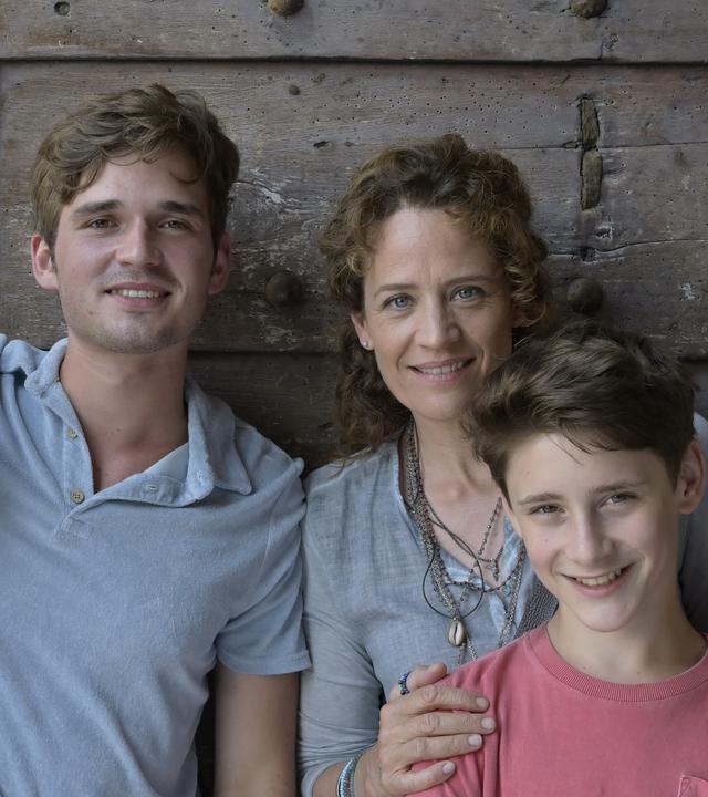 "Ein Sommer auf Elba": Vor einer dunklen Tür auf einer Terrasse steht Maja und ihre Familie. Von links: ihr Ex-Mann Thorsten (Janek Rieke), daneben ihr 17-jähriger Sohn Anton (Henry Horn). Maja (Regula Grauwiller) hat ihrem jüngeren Sohn Erik (Levis Kachel) liebevoll die Hände auf die Schultern gelegt.