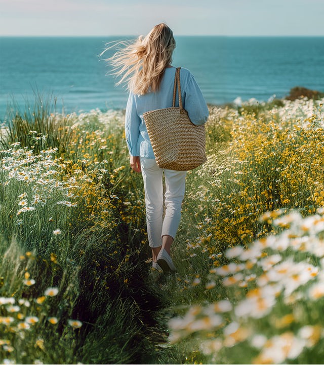 Eine Frau läuft in einer verträumten Blumenlanschaft von der betrachtenden Person Weg. Am Horizont ist das Meer zu erkennen. Die Frau trägt eine blaue Bluse, eine weiße Hose und eine große Strandtasche.