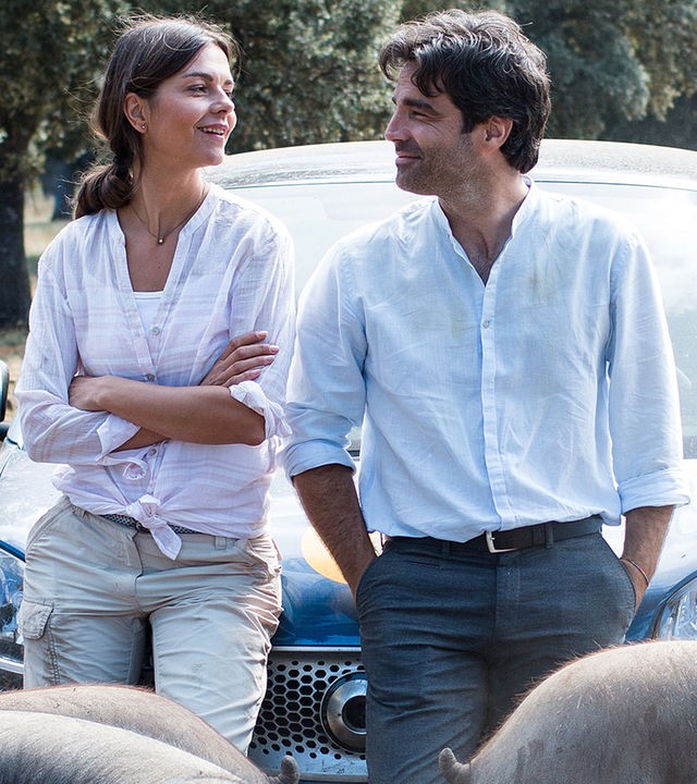 Carlos (Fernando Corral) und Rike (Susan Hoecke), umringt von vielen Schweinen, lehnen auf einer Motorhaube und schauen sich lächelnd an.