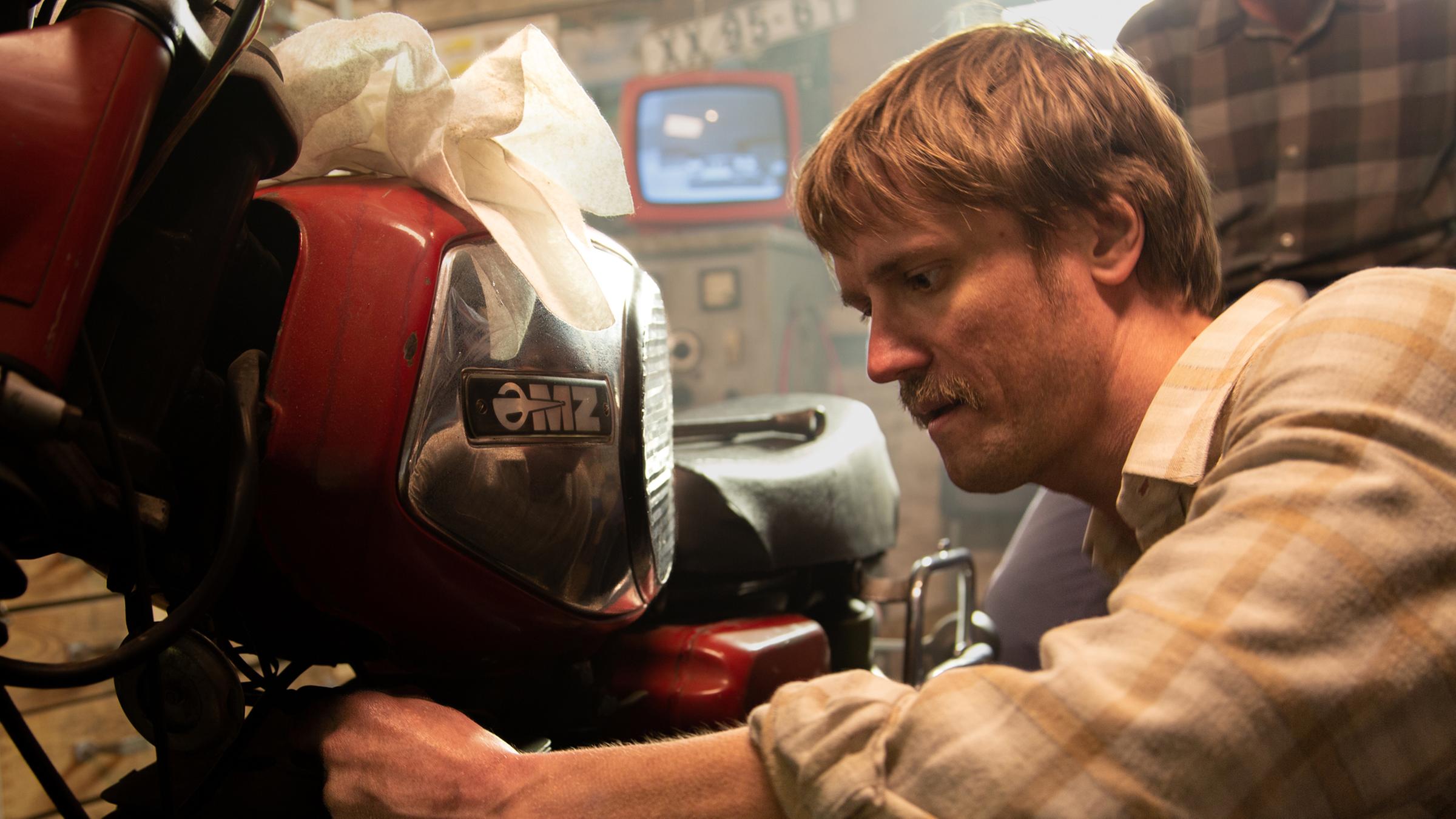 Hans Joachim Zorn (Florian Donath) schraubt an seinem Motorrad, im Hintergrund ein Fernseher und ein weiterer Mann.