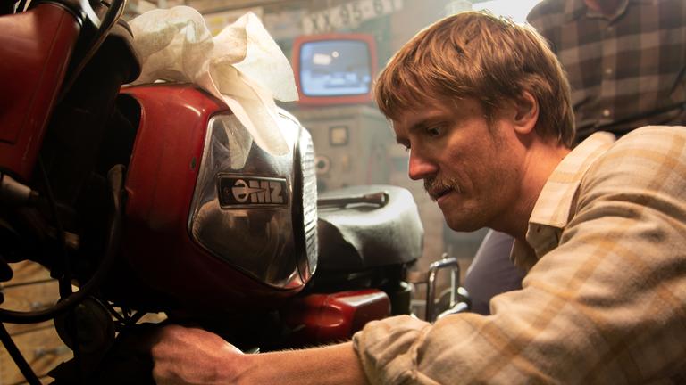 Hans Joachim Zorn (Florian Donath) schraubt an seinem Motorrad, im Hintergrund ein Fernseher und ein weiterer Mann.