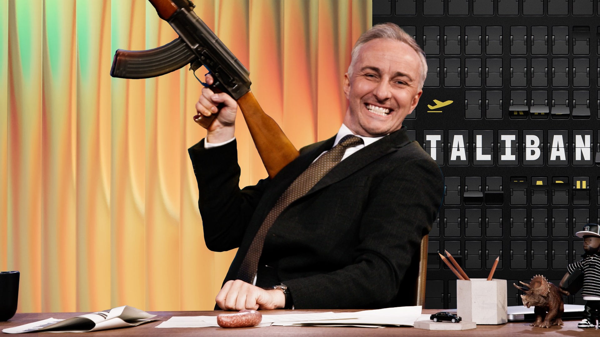 Jan Böhmermann sitzt mit breitem Grinsen am Schreibtisch. Er hält eine Kalaschnikow schräg nach oben gerichtet. Rechts im Hintergrund ist eine Anzeigetafel mit einem kleinen gelben Flugzeug abgebildet, in der Mitte der Tafel steht: TALIBAN.