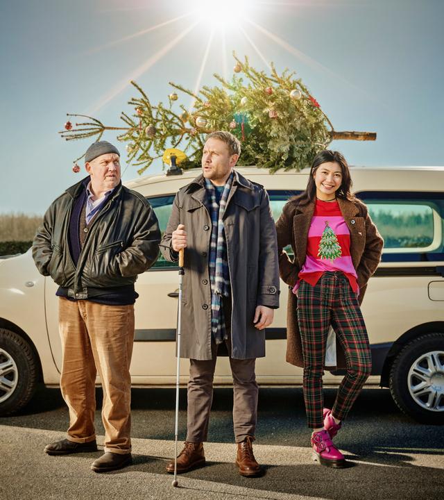 Drei Personen stehen vor einem hellen Kombi, auf dessen Dach ein geschmückter Weihnachtsbaum befestigt ist – bereit für die festliche Fahrt.