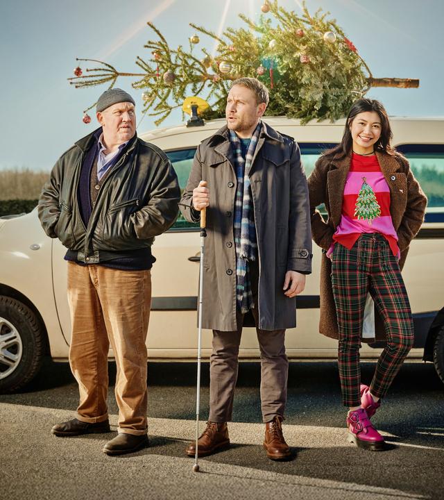 Drei Personen stehen vor einem hellen Kombi, auf dessen Dach ein geschmückter Weihnachtsbaum befestigt ist – bereit für die festliche Fahrt.