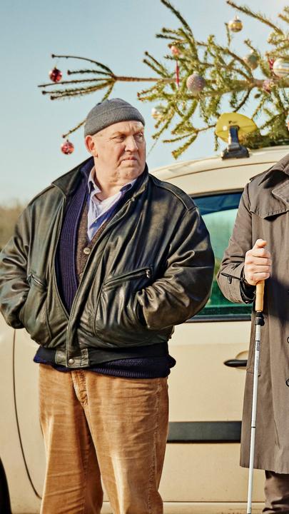 "Ein Taxi zur Bescherung": Axel (Dietmar Bär), Jan (Max Riemelt) und Linh (Nhung Hong) stehen nebeneinander vor einem Taxi, auf dessen Dach ein mit Kugeln geschmückter Tannenbaum festgebunden ist. Jan hält seinen Taststock in der Hand. Die Sonne scheint vom winterlich-blauen Himmel.