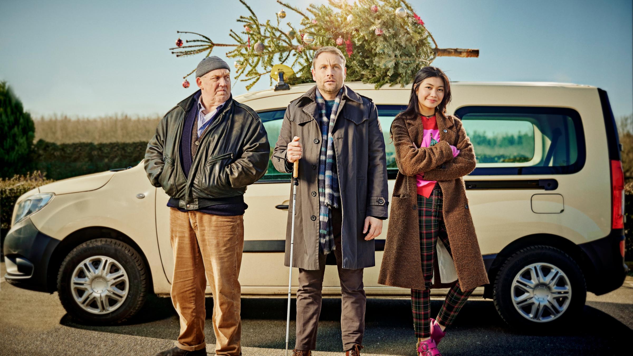 "Ein Taxi zur Bescherung": Axel (Dietmar Bär), Jan (Max Riemelt) und Linh (Nhung Hong) stehen nebeneinander vor einem Taxi, auf dessen Dach ein mit Kugeln geschmückter Tannenbaum festgebunden ist. Jan hält seinen Taststock in der Hand. Die Sonne scheint vom winterlich-blauen Himmel.