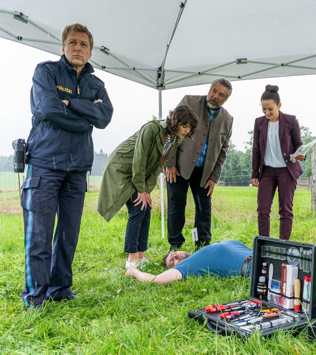 "Die Rosenheim-Cops - Ein überraschendes Wiedersehen": Polizeihauptmeister Mohr (Max Müller), Kommissarin Andresen (Sophie Melbinger), Kommissar Stadler (Dieter Fischer), Pathologin Atay (Sevda Polat) und der Spurensicherer (Komparse) stehen auf einer Wiese um das Opfer (Komparse) herum, das auf dem Rücken liegt. Im Vordergrund ist ein Koffer mit Utensilien zur Spurensicherung zu sehen.