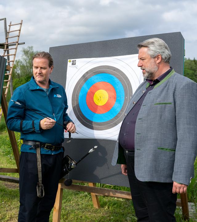 "Die Rosenheim-Cops - Ein Unfall mit Folgen": Die Kommissare Hansen (Igor Jeftić) und Stadler (Dieter Fischer) sprechen mit dem Bogenschützen Walter Hofmeister (Martin Dudeck), der mit einem Pfeil in der Hand vor einer Zielscheibe auf einer Wiese steht.