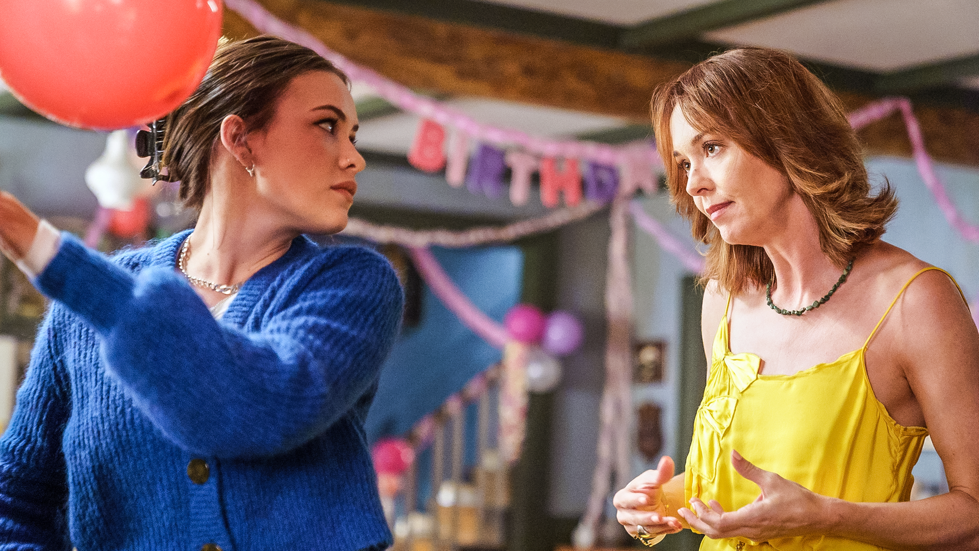 Issie und ihre Mutter Amanda sehen einander genervt an. Issie hält einen Ballon in der Hand, im Hintergrund hängt eine "Happy Birthday"-Girlande.