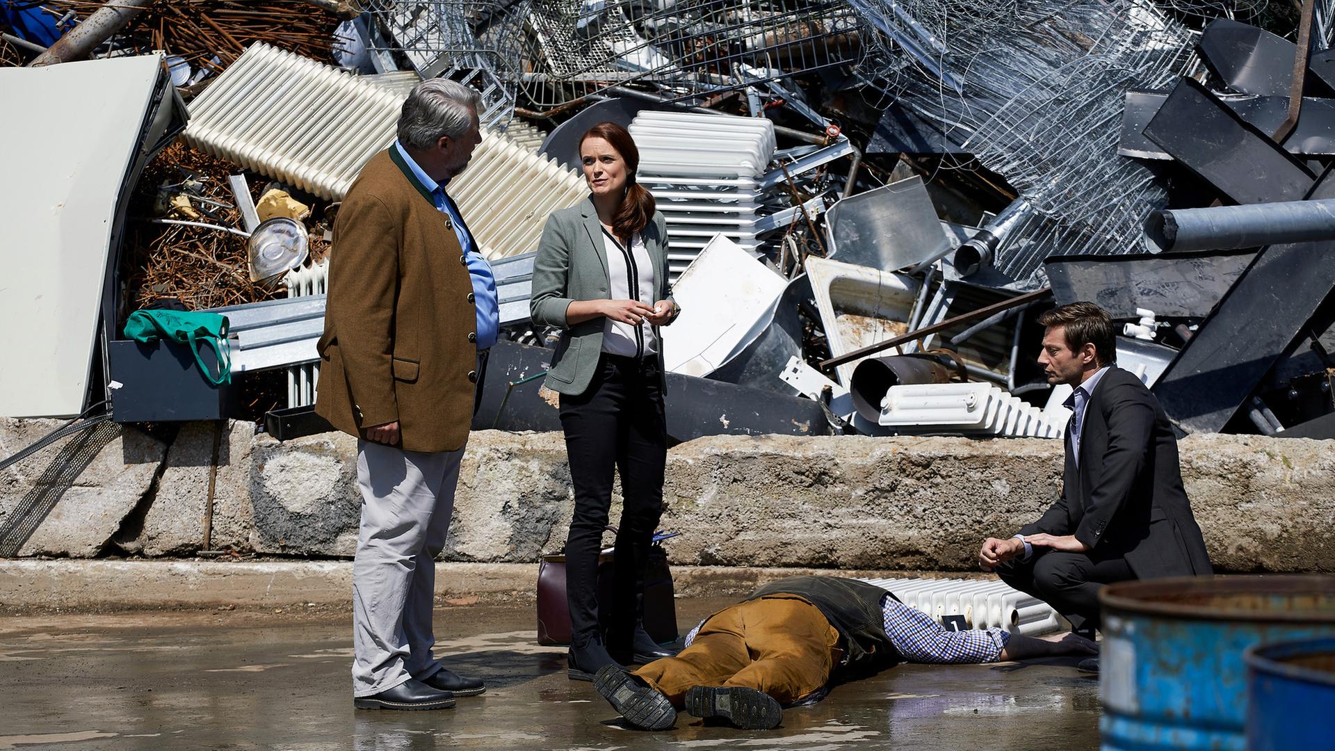 "Die Rosenheim-Cops - Ein Vater kommt selten allein": Kommissar Stadler (Dieter Fischer), Sandra Mai (Sina Wilke) und Kommissar Hansen (Igor Jeftic) bilden einen Halbkreis um das Mordopfer, dass bäuchlings auf dem Asphalt vor einem Berg Altmetall liegt. Vor dem Kopf des Opfers liegt ein Heizkörper.