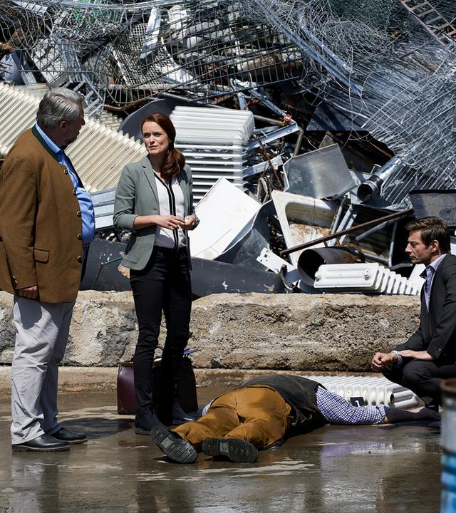 "Die Rosenheim-Cops - Ein Vater kommt selten allein": Kommissar Stadler (Dieter Fischer), Sandra Mai (Sina Wilke) und Kommissar Hansen (Igor Jeftic) bilden einen Halbkreis um das Mordopfer, dass bäuchlings auf dem Asphalt vor einem Berg Altmetall liegt. Vor dem Kopf des Opfers liegt ein Heizkörper.
