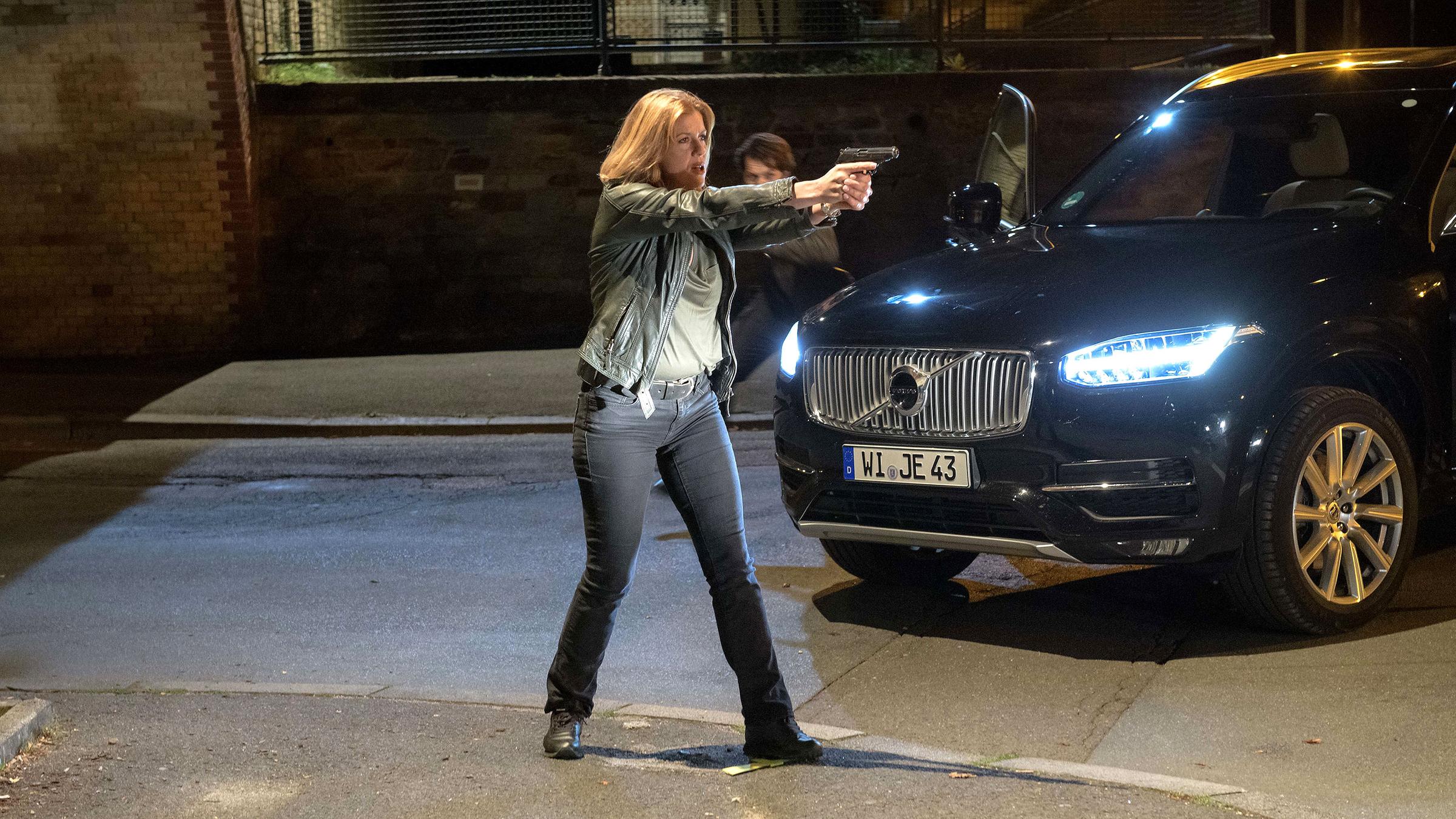 Kerstin Klar (Fiona Coors) steht im Scheinwerferlicht eines schwarzen Autos auf der Straße. Mit ausgestreckten Armen hält sie ihre Waffe vor sich. Im Hintergrund erkennt man Nikos Weber (Jeff Wilbusch), der sich hinter dem Wagen befindet.