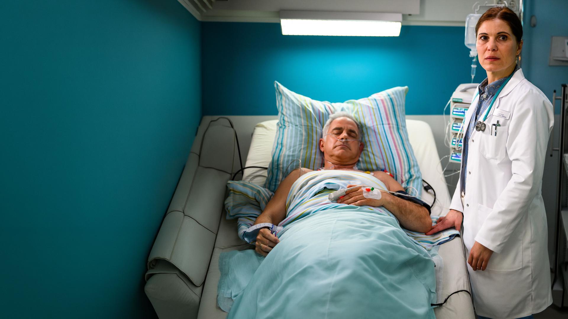 "Bettys Diagnose - Einbrüche": Prof. Dr. Alexander von Arnstett (Florian Fitz) liegt mit geschlossenen Augen im Krankenbett. Ein Sauerstoffschlauch führt in seine Nase. Neben seinem Bett steht Dr. Nadira Abbas (Yasmina Djaballah), die besorgt in die Ferne schaut.