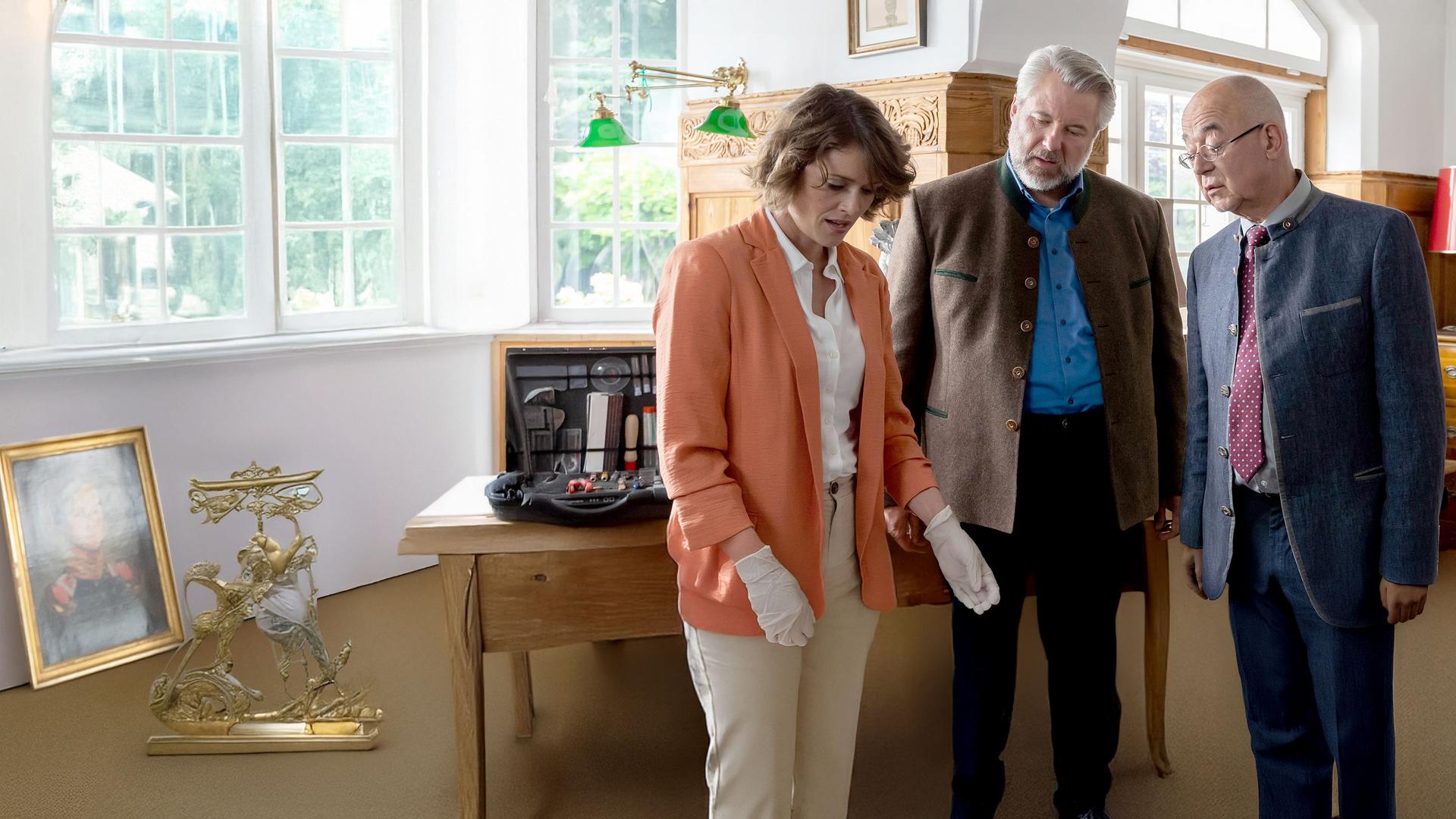 Kommissar Stadler und Polizeidirektor Achtziger stehen am Schreibtisch des Opfers, das neben ihnen auf dem Boden liegt, und schauen auf ein bebildertes Dokument, in dem Andresen blättert.