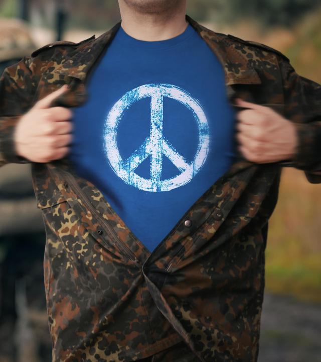 Ein Mann in Flecktarn reißt seine Jacke auf und trägt darunter ein T-Shirt mit einem Peace-Zeichen.