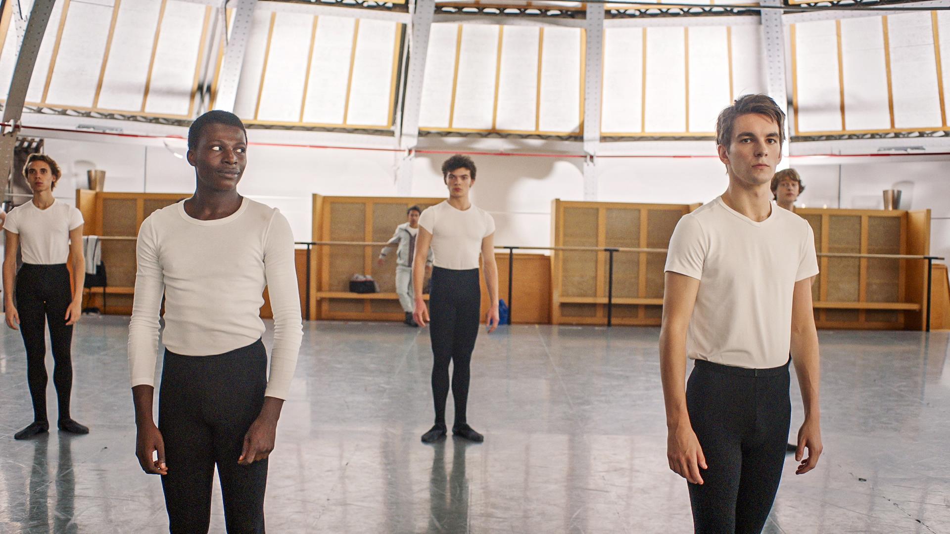 Isaac (links) und Max (rechts) proben für den Grand Prix. Im Hintergrund sind ihre Mitschüler zu sehen. Die beiden sehen ernst aus und Isaac schaut zu Max rüber.