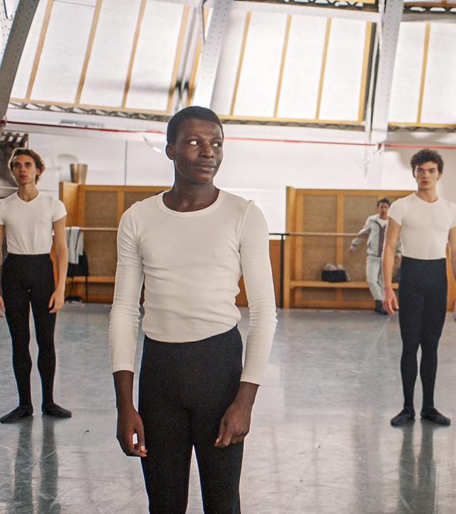 Isaac (links) und Max (rechts) proben für den Grand Prix. Im Hintergrund sind ihre Mitschüler zu sehen. Die beiden sehen ernst aus und Isaac schaut zu Max rüber.