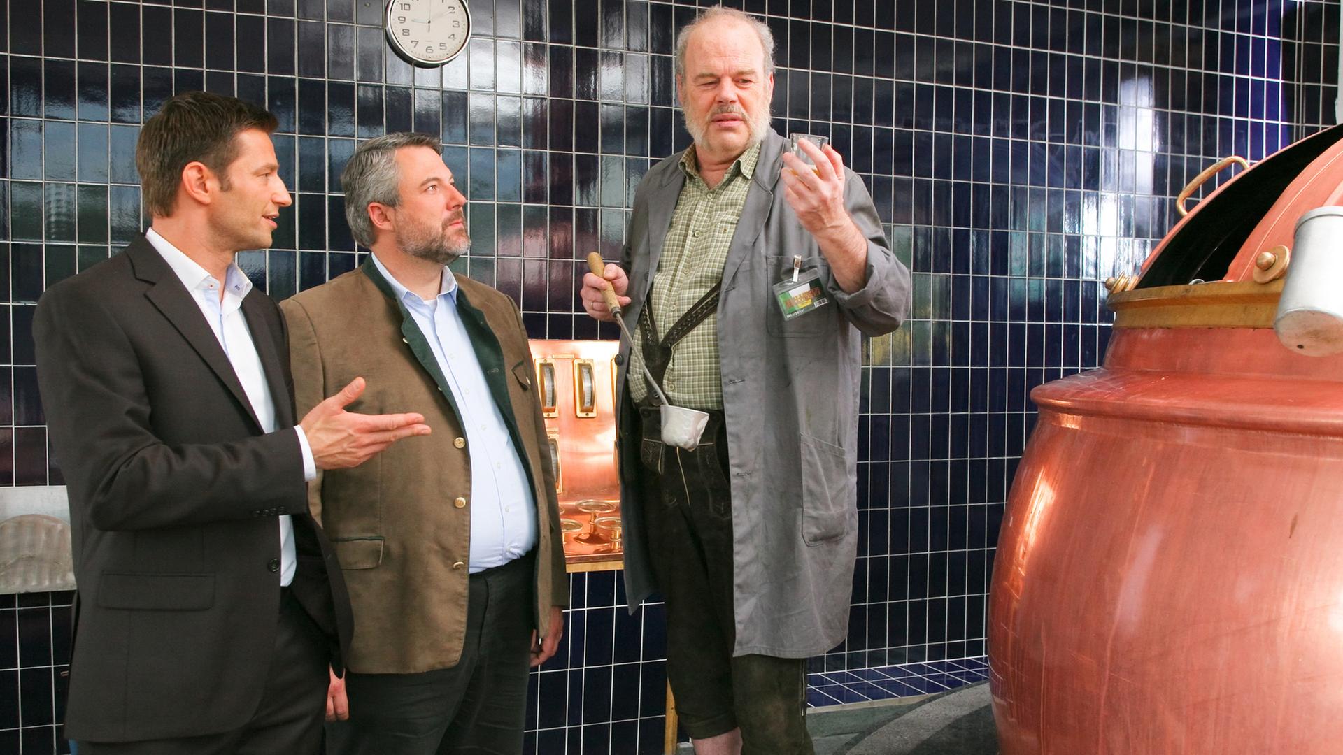 Die Kommissare Hansen (Igor Jeftic) und Stadler (Dieter Fischer) stehen neben Josef Moosbach (Andreas Pegler), der ein Glas und eine Kelle in den Händen hält. Er steht neben einem großen Kupferkessel.