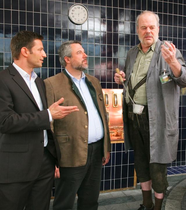 Die Kommissare Hansen (Igor Jeftic) und Stadler (Dieter Fischer) stehen neben Josef Moosbach (Andreas Pegler), der ein Glas und eine Kelle in den Händen hält. Er steht neben einem großen Kupferkessel.