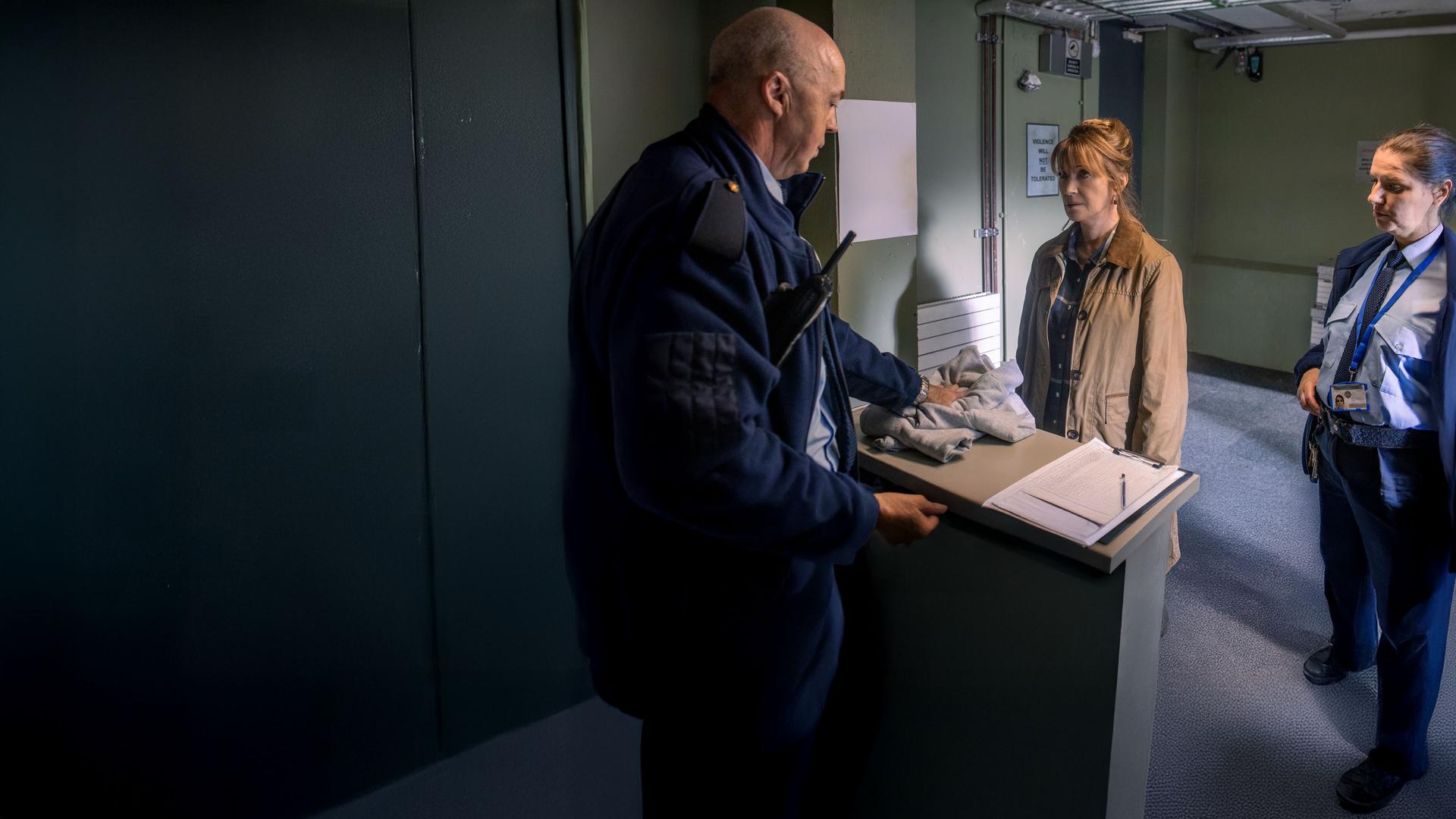 "Harry Wild - Mörderjagd in Dublin: Eine Leiche zum Dessert": Harry Wild (Jane Seymour) steht in der Aufnahme des Gefängnisses. Der ihr gegenüberstehende, uniformierte Polizist mit Funkgerät in der rechten Brusttasche, überreicht ihr die Gefängniswäsche. Neben Harry steht eine uniformierte Polizistin.